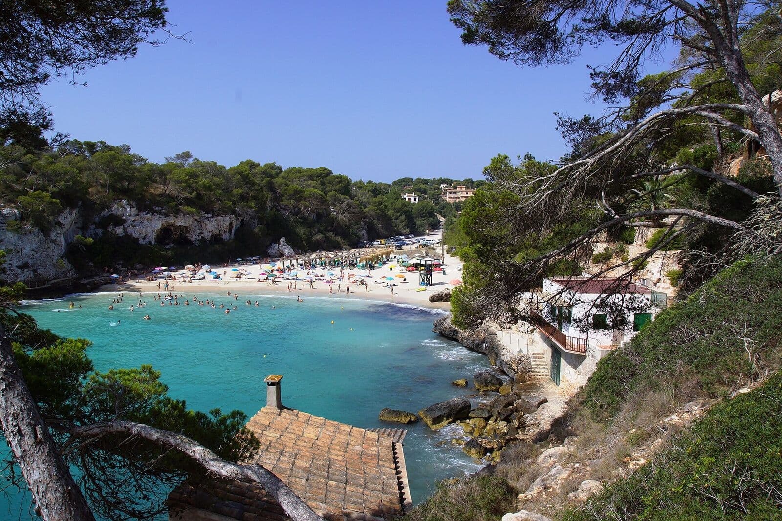 Cala Llombards vista do alto, com areia branca, água turquesa, pinheiros e o telhado de um abrigo de barcos em primeiro plano