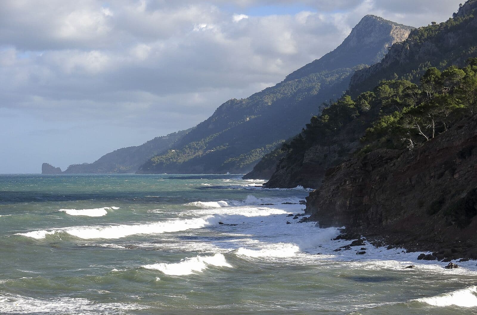 Costa rochosa de Port des Canonge com ondas, pinheirais e as montanhas da Tramuntana ao fundo