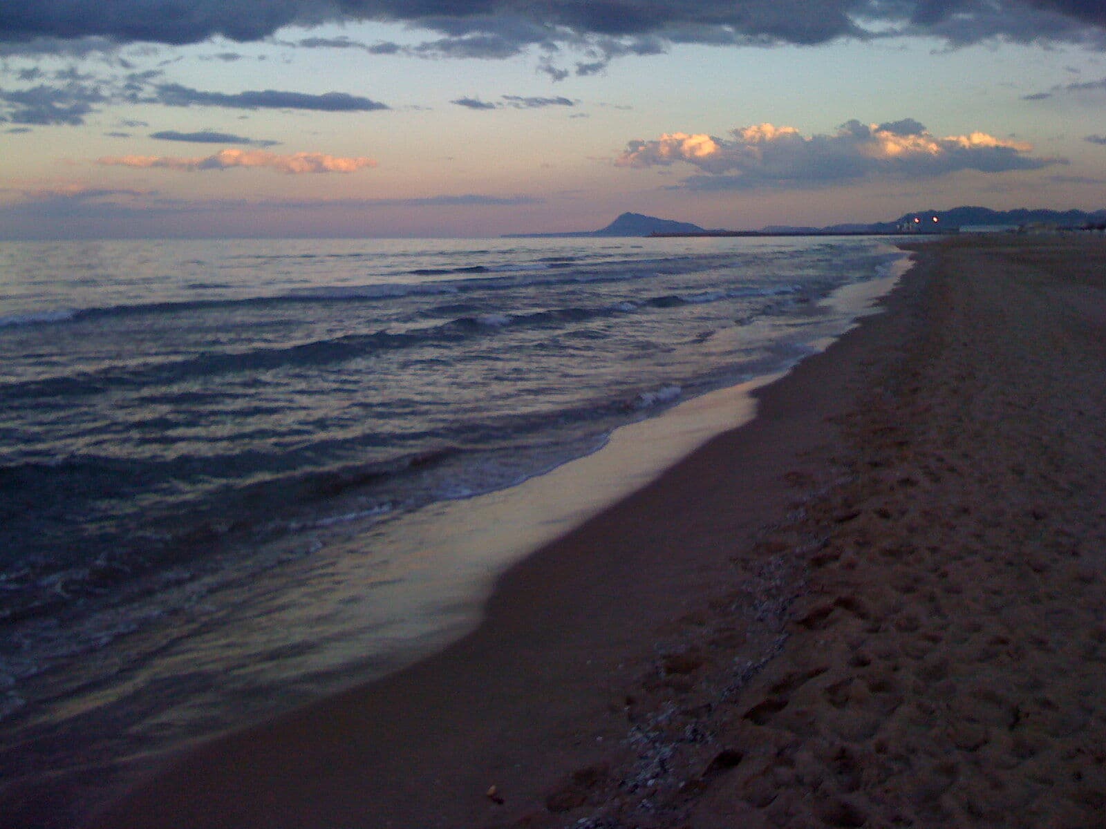 The shoreline of Playa de Gandia at dusk, with gentle waves, wet sand and mountains in the distance under a cloudy sky