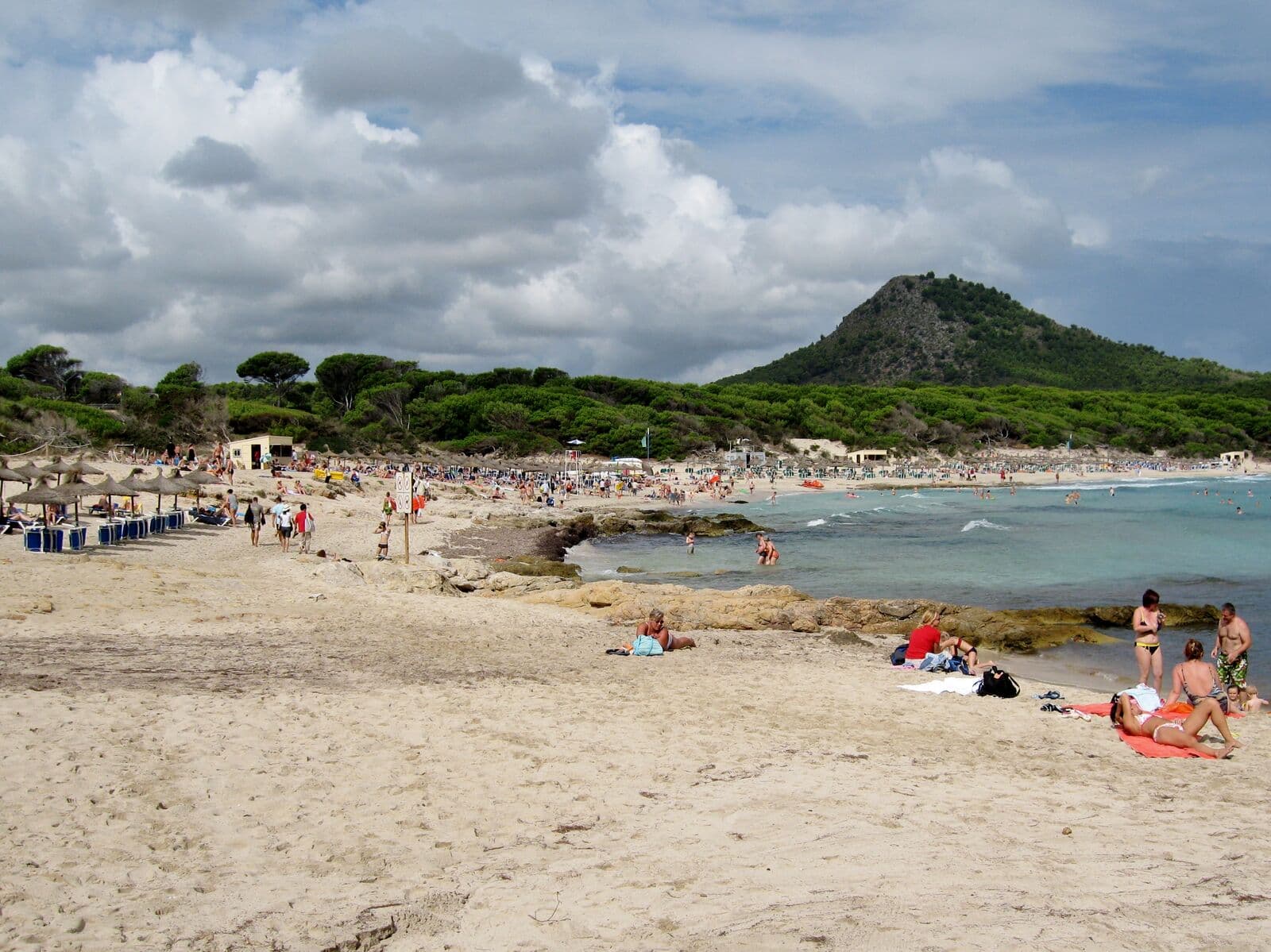 Bathers on the pale sand of Cala Agulla, with pine woods and a green hill behind the turquoise water