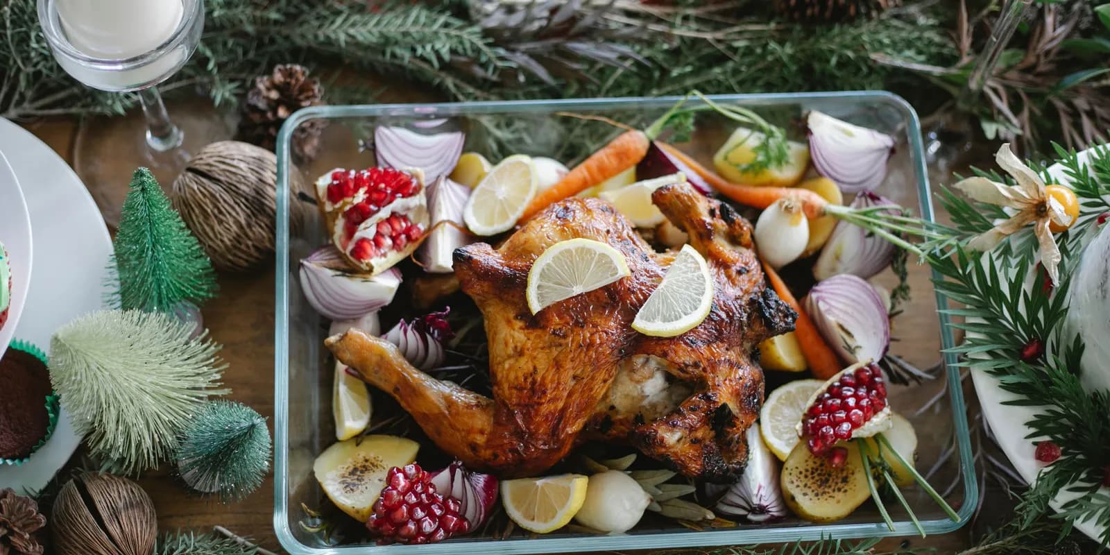 Whole roast chicken in a glass dish with red onion, carrots, potatoes, lemon slices and pomegranate, on a Christmas table with fir branches.
