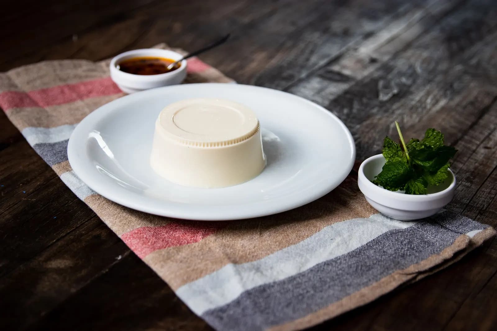 White panna cotta turned out onto a plate on a striped cloth, with a small jug of caramel and a bowl of mint.