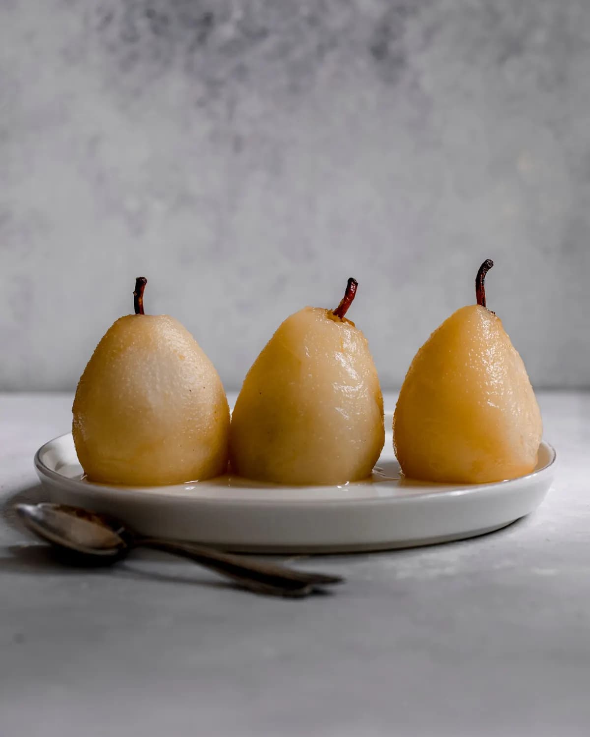 Three whole poached pears, peeled with stalks on, standing on a white plate with syrup, next to two teaspoons.