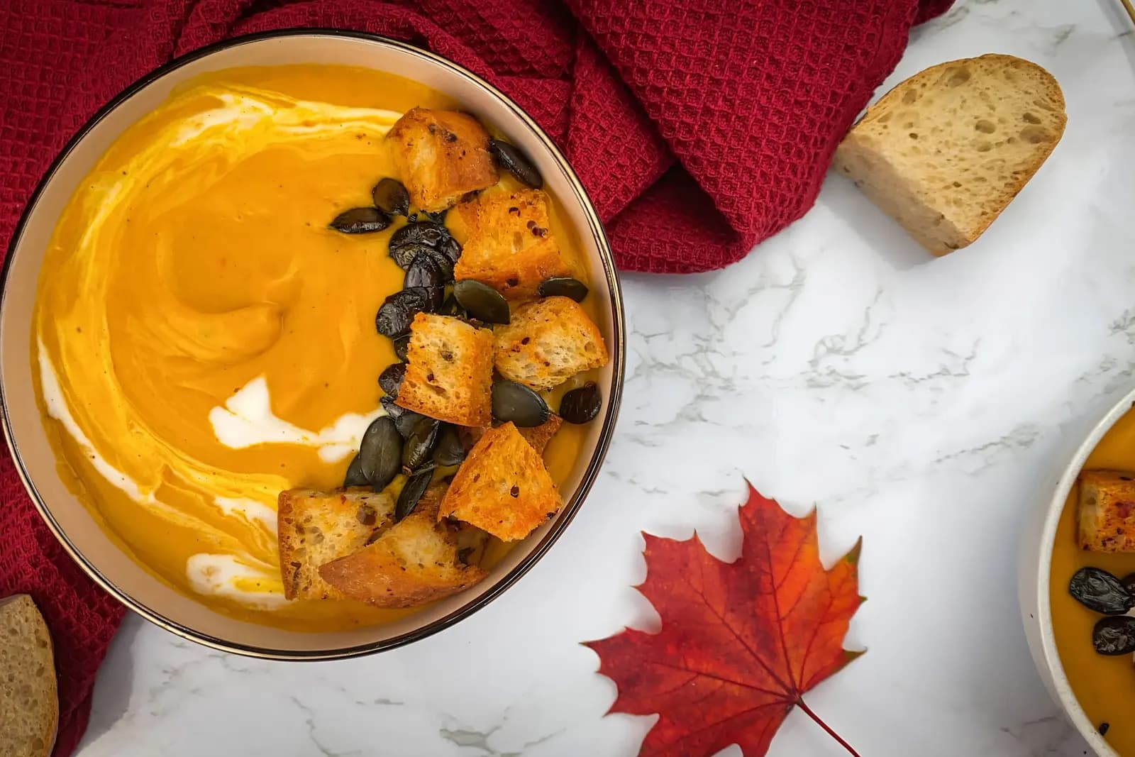 Bowl of orange pumpkin soup with croutons, pumpkin seeds and a swirl of cream, next to a red cloth and bread.