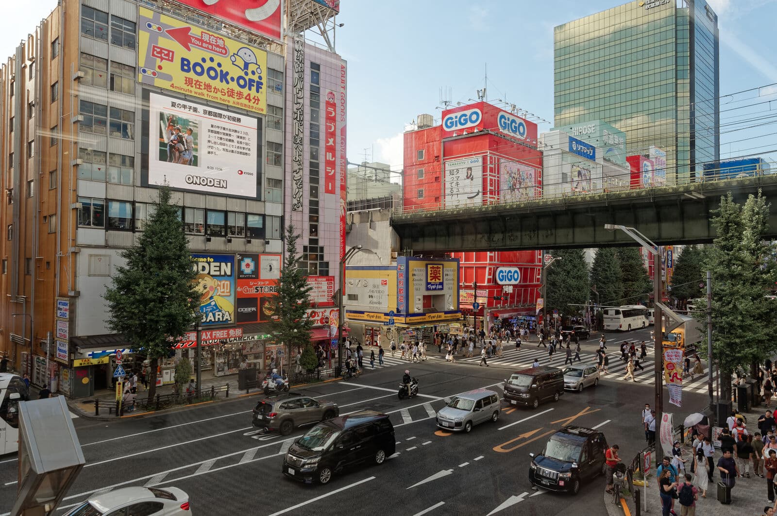 Un carrefour à Akihabara, Tokyo, avec des voitures, des piétons sur le passage, des immeubles couverts d'enseignes publicitaires et un pont ferroviaire en arrière-plan