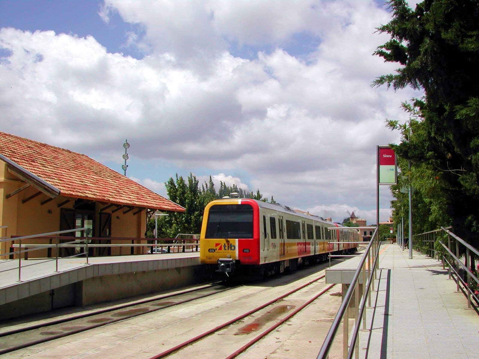 Trem da SFM parado na plataforma da estação de Sineu, ao lado de um prédio com telhado vermelho