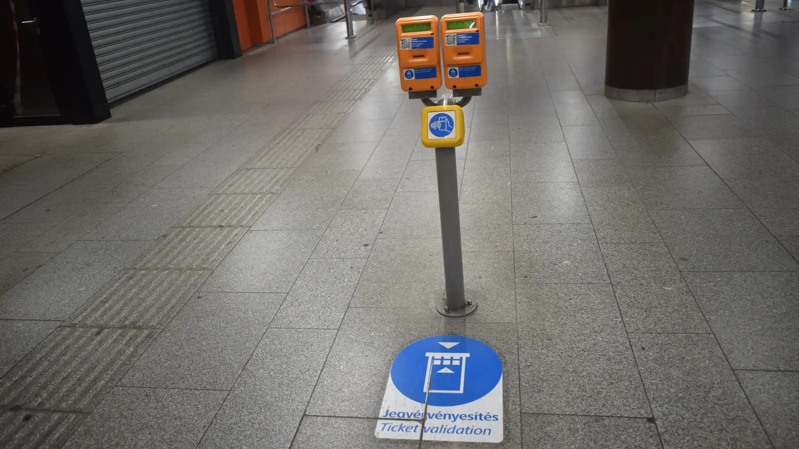 Composteurs de billets orange sur un poteau dans le hall d’une station de métro, avec un panneau bleu au sol indiquant où valider.