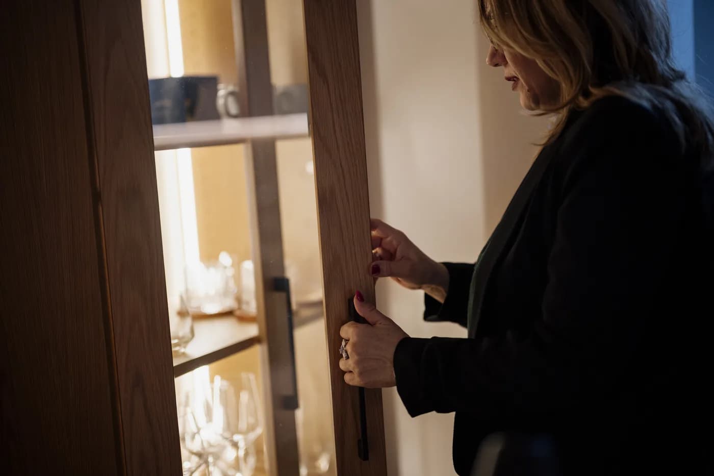 A person opens a wooden kitchen cabinet to reach glassware on illuminated shelves; this is an ordinary cabinet door, not a pocket-door kitchen system.