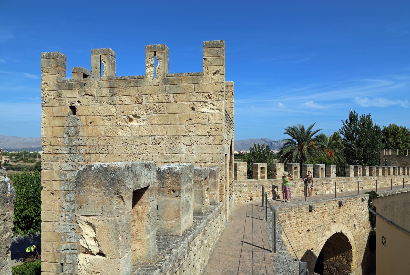 Passeio sobre a muralha de Alcúdia, com ameias de pedra, uma torre e palmeiras