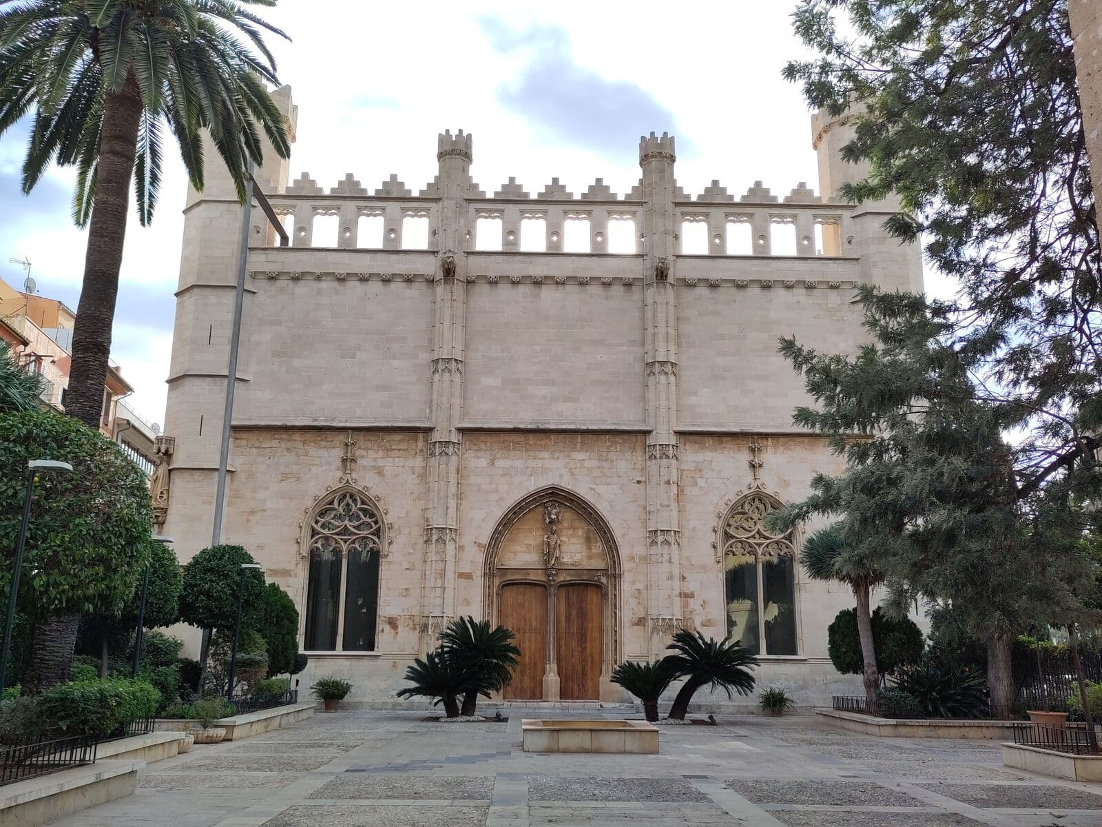 Fachada gótica da Llotja de Palma, com ameias, janelões e uma grande porta, vista de um jardim com palmeiras