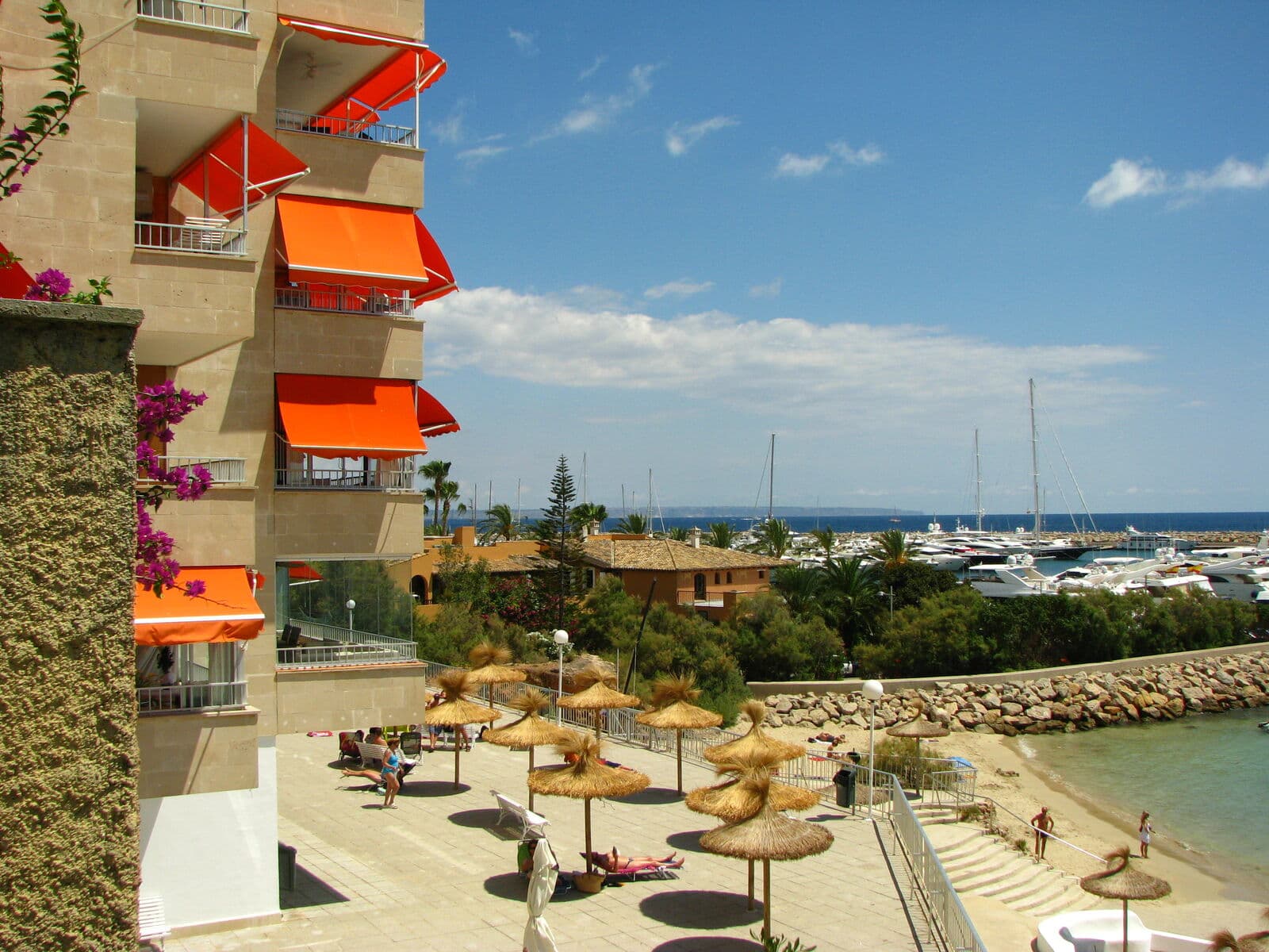 Calçadão junto a uma pequena praia em Portals Nous, com prédios de apartamentos, guarda-sóis de palha e os barcos de Puerto Portals ao fundo