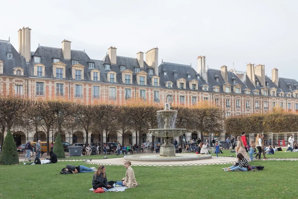 Brick and stone façades of the Place des Vosges, with people relaxing on the lawn and a fountain.