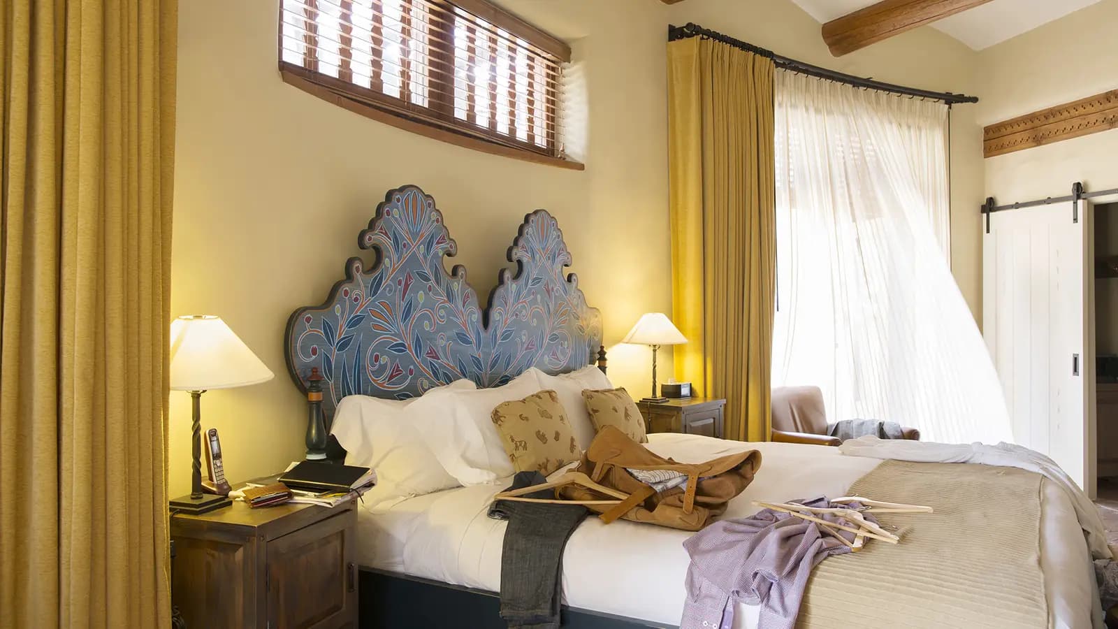 Habitación de hotel con cama doble, cabecero pintado en azul, lámparas encendidas y ropa sobre la colcha junto a una ventana con cortinas.