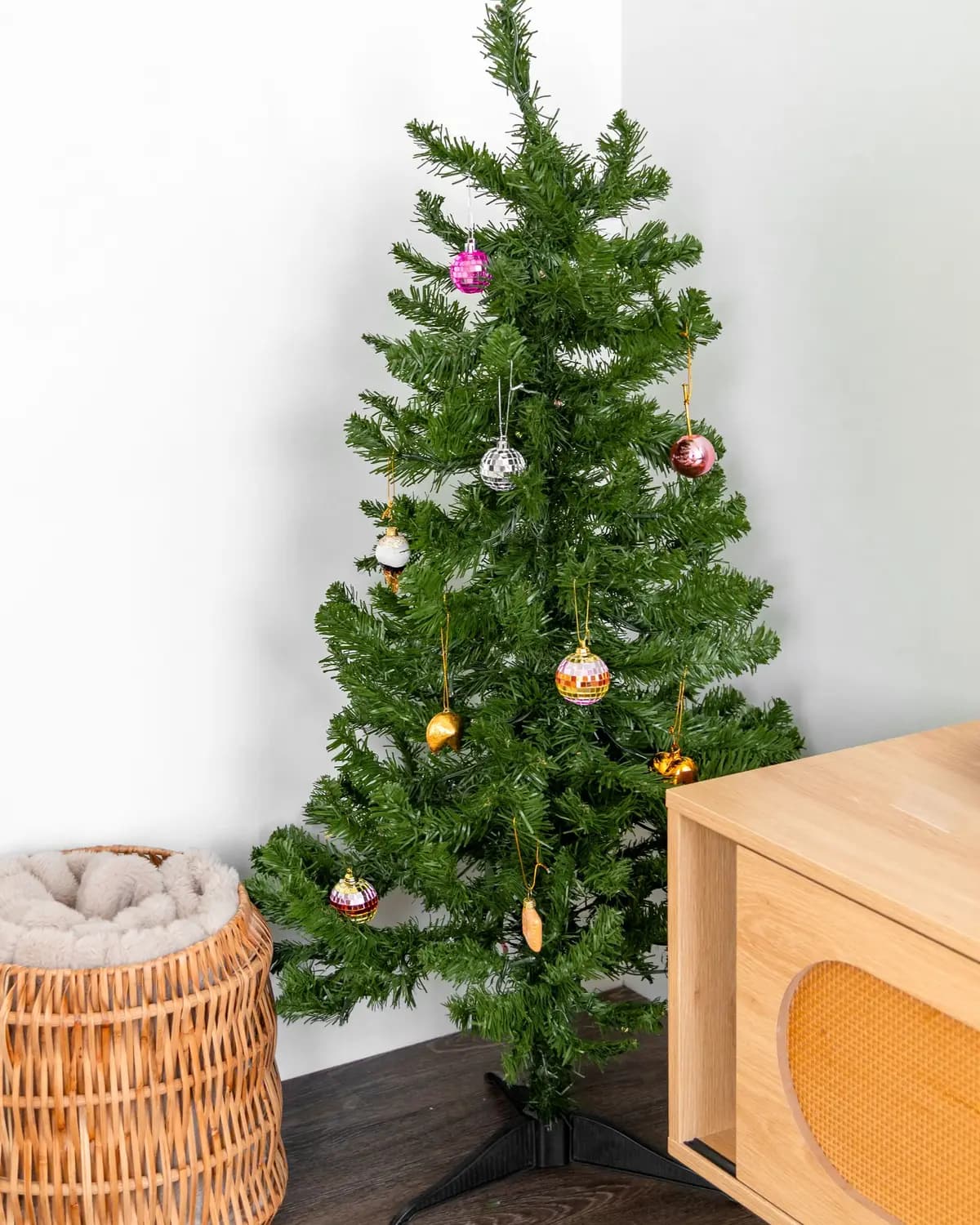 Pequena árvore de Natal artificial no canto de uma divisão, com poucas bolas coloridas, ao lado de um cesto de vime e de um móvel de madeira clara.