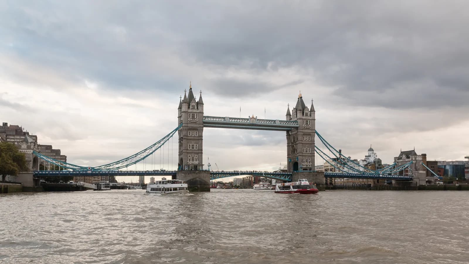 Tower Bridge au-dessus de la Tamise sous un ciel nuageux, avec deux bateaux touristiques sur le fleuve.