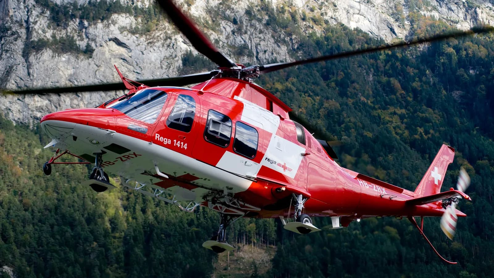 Rot-weißer Rettungshubschrauber startet neben einem bewaldeten Berghang in den Schweizer Alpen.